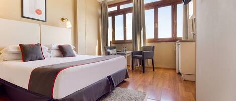 Une chambre d’hôtel avec un grand lit, un bureau et une chaise. Il y a une fenêtre avec des rideaux, un tableau encadré au mur et un radiateur.