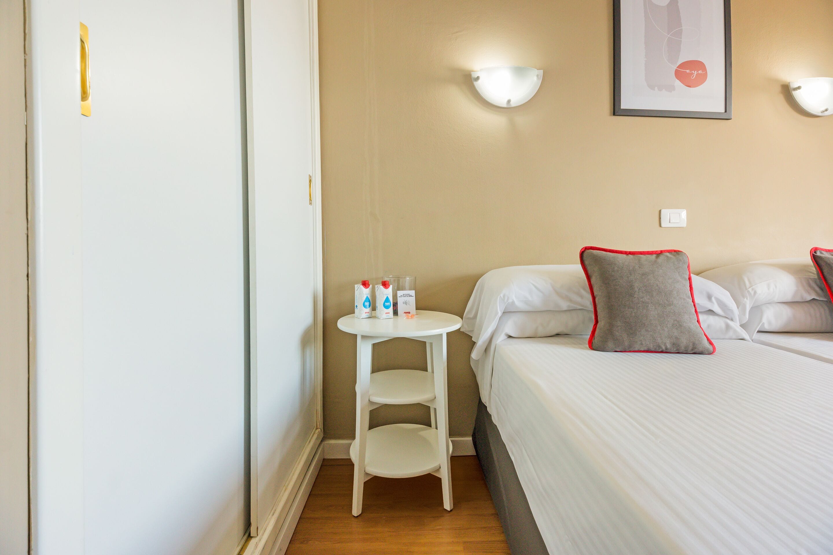 A hotel room with a bed, a small white table with toiletries, a wall-mounted light, and a framed picture.