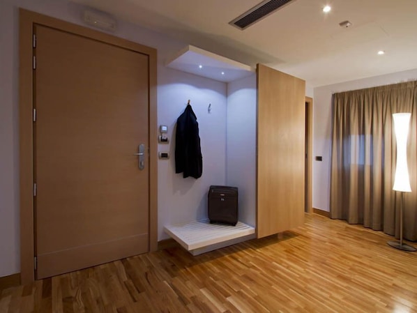 Una camera d'albergo con pavimento in legno, una porta chiusa, una panca con una valigia, un appendiabiti e una lampada.