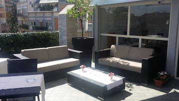Un patio exterior con mobiliario de mimbre, incluyendo sofás y una mesa de centro, y una pared de vidrio que ofrece vistas de los edificios circundantes.