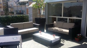 An outdoor patio area with wicker furniture, including sofas and a coffee table, and a glass wall offering a view of the surrounding buildings. - Hotel Gravina San Pietro (Rome)