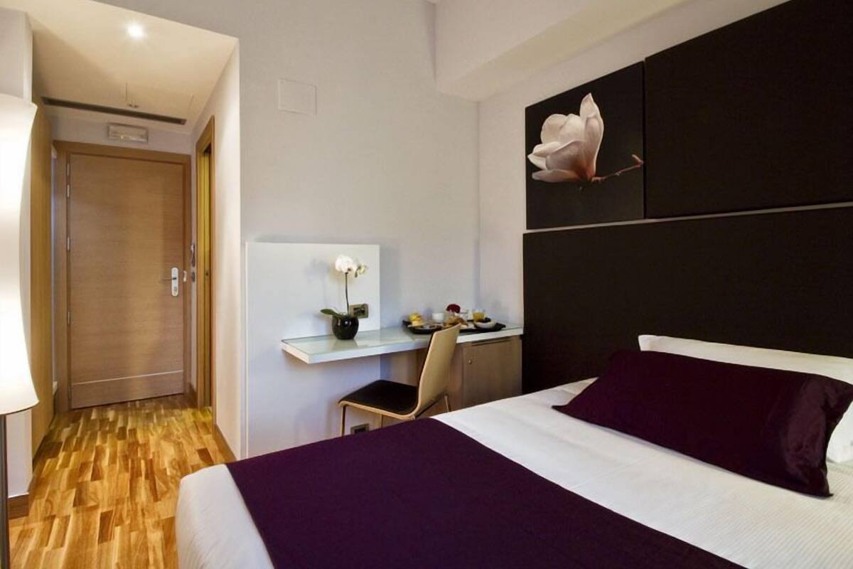 Hotellihuone, jossa on sänky, työpöytä tuolilla, maljakko kukalla ja seinälle ripustettu taideteos.