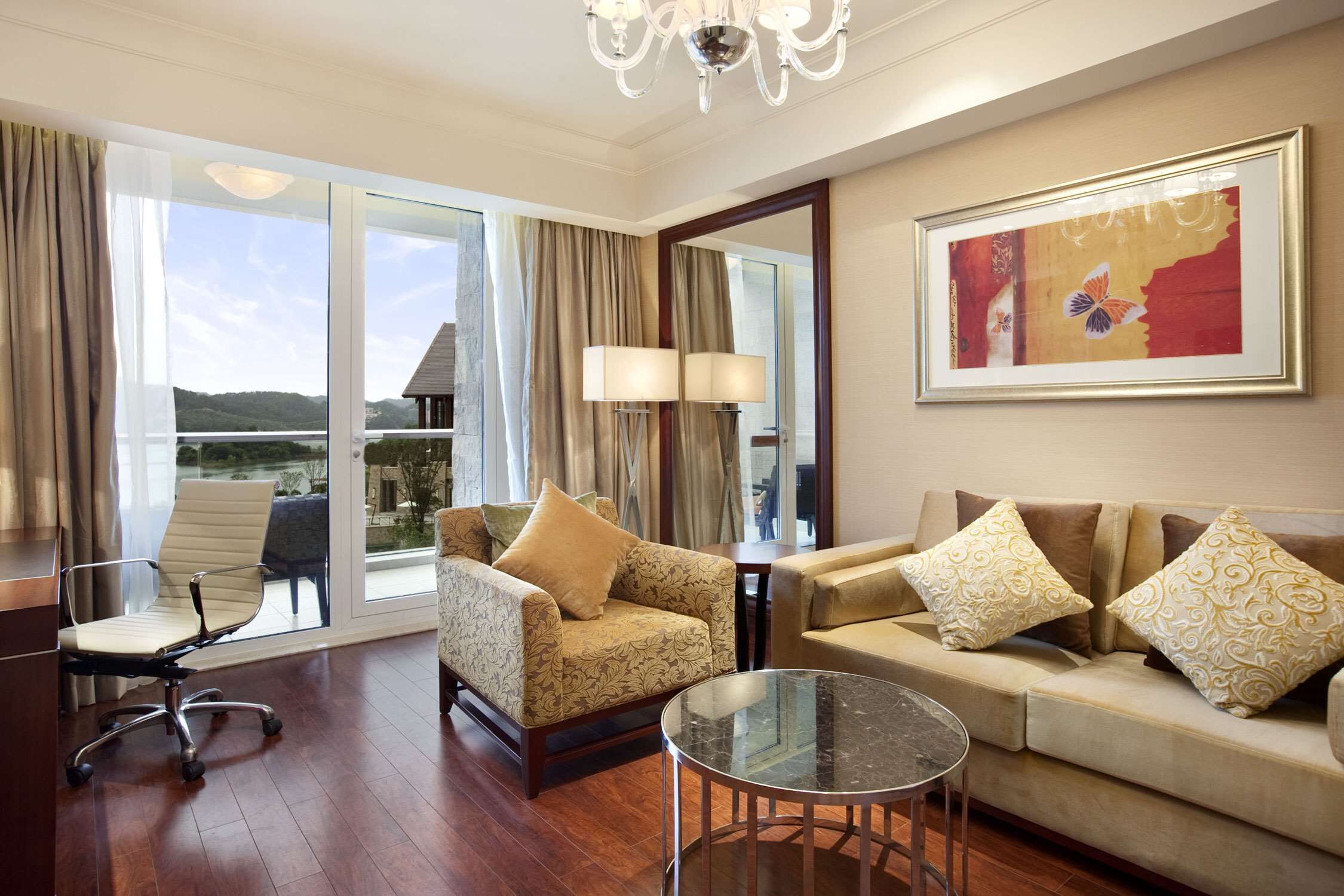 A spacious living room with a sofa, armchair, and glass coffee table. A large window offers a view of the outdoors.