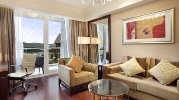 A spacious living room with a sofa, armchair, and glass coffee table. A large window offers a view of the outdoors.