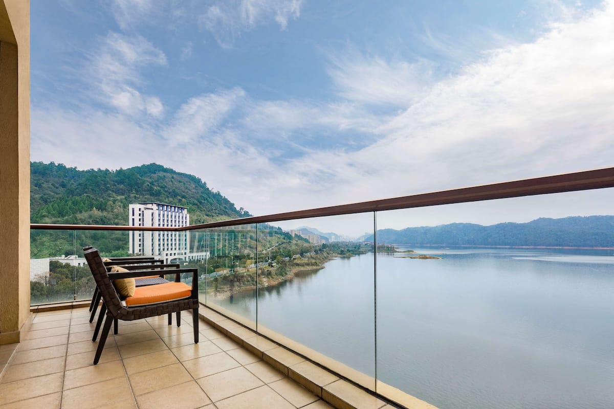 A balcony with a glass railing overlooking a river, a chair with an orange cushion, and a building in the background.