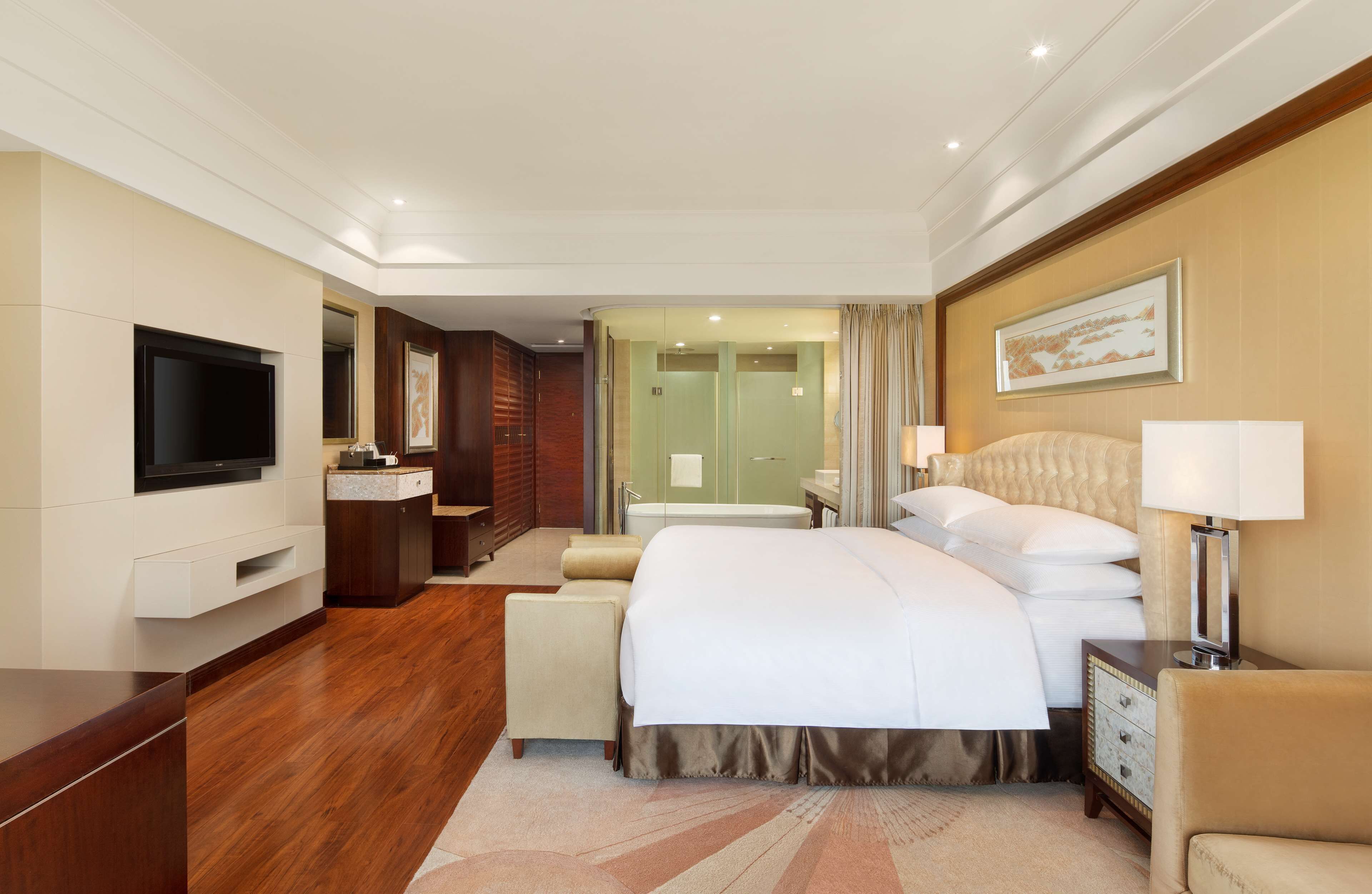Une chambre d’hôtel avec un grand lit, un téléviseur à écran plat, une coiffeuse avec un miroir, une salle de bain avec une baignoire et un tableau encadré au mur.