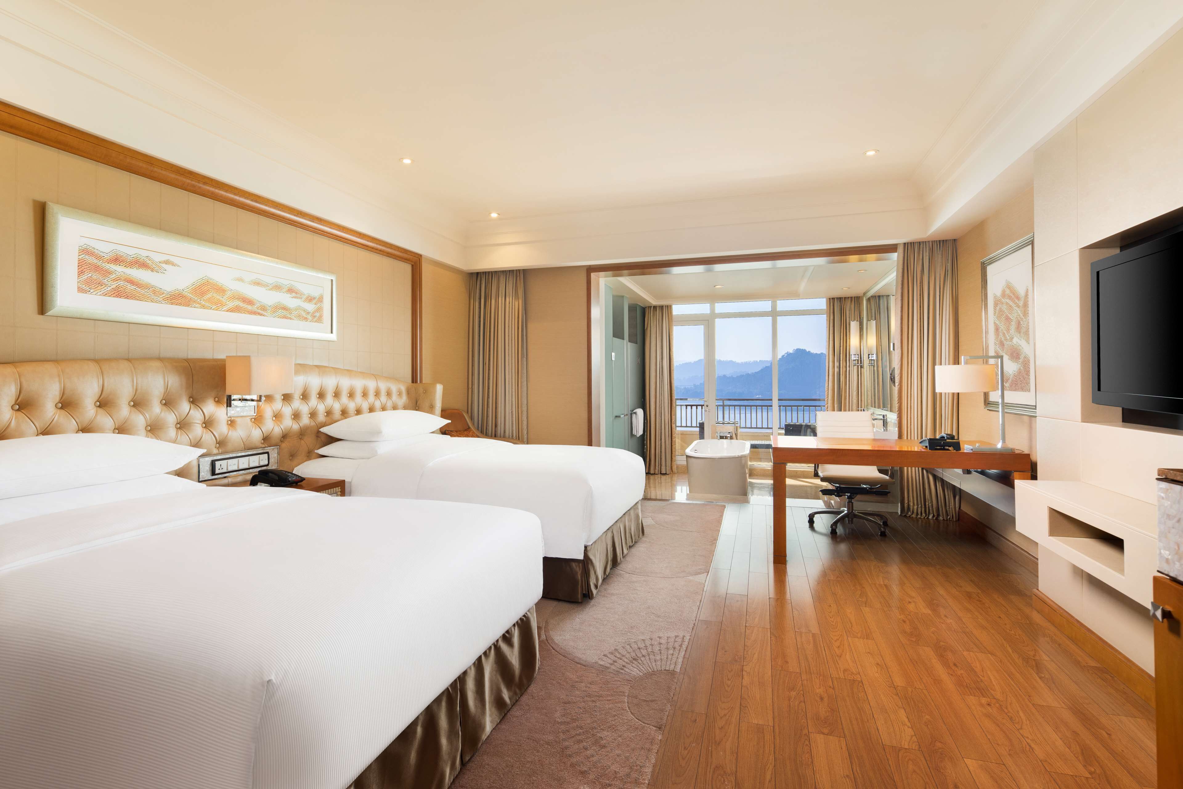 A hotel room with two beds, a desk, a TV, and a balcony with a mountain view.