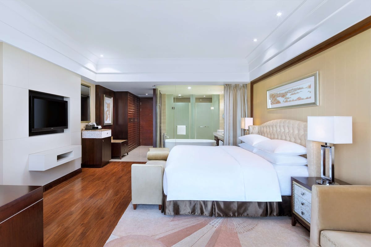 A modern hotel room with a large bed, a flat-screen TV, a vanity with a mirror, a bathroom with a glass partition, and a framed artwork on the wall.