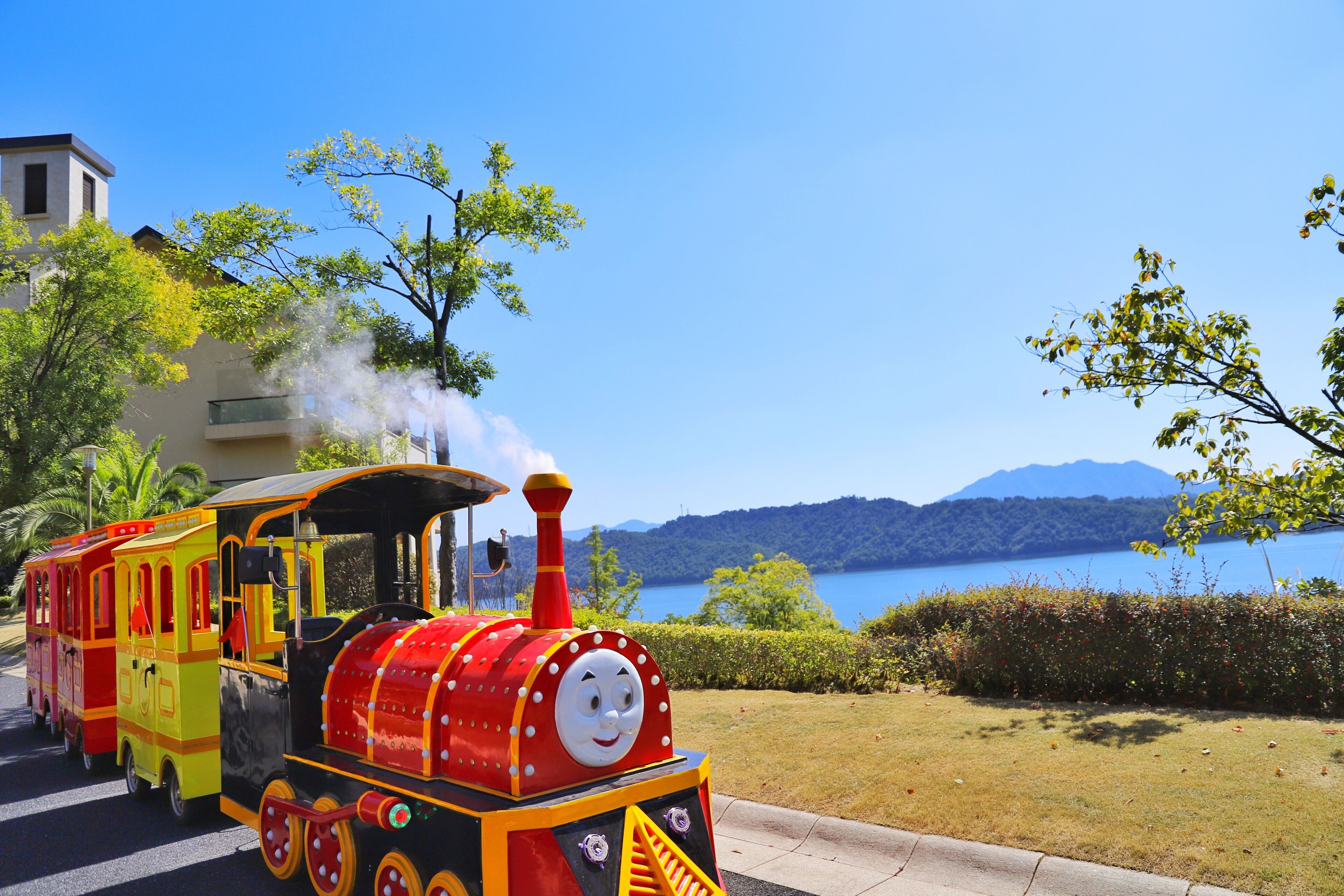 A colorful train with a smiling face, moving along a track with a scenic lake and mountains in the background.
