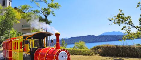 A colorful train with a smiling face, moving along a track with a scenic lake and mountains in the background.