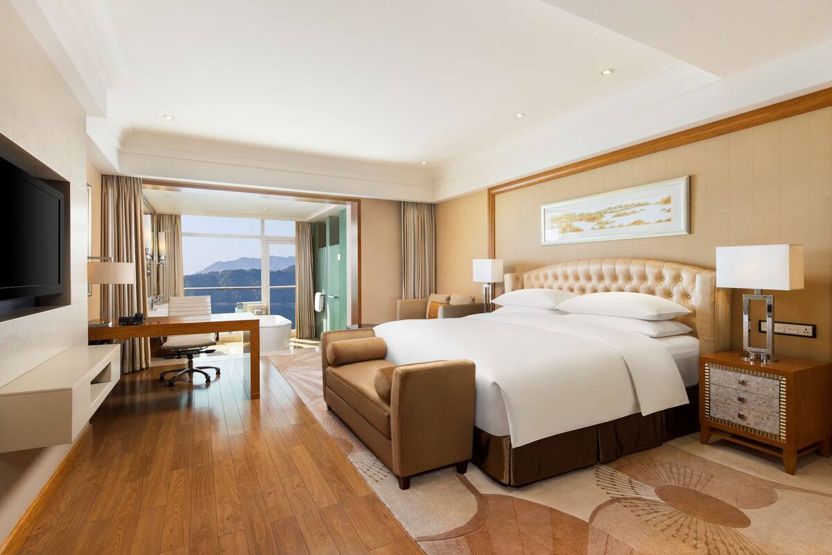A hotel room with a large bed, a desk, a chair, a TV, and a view of the mountains.