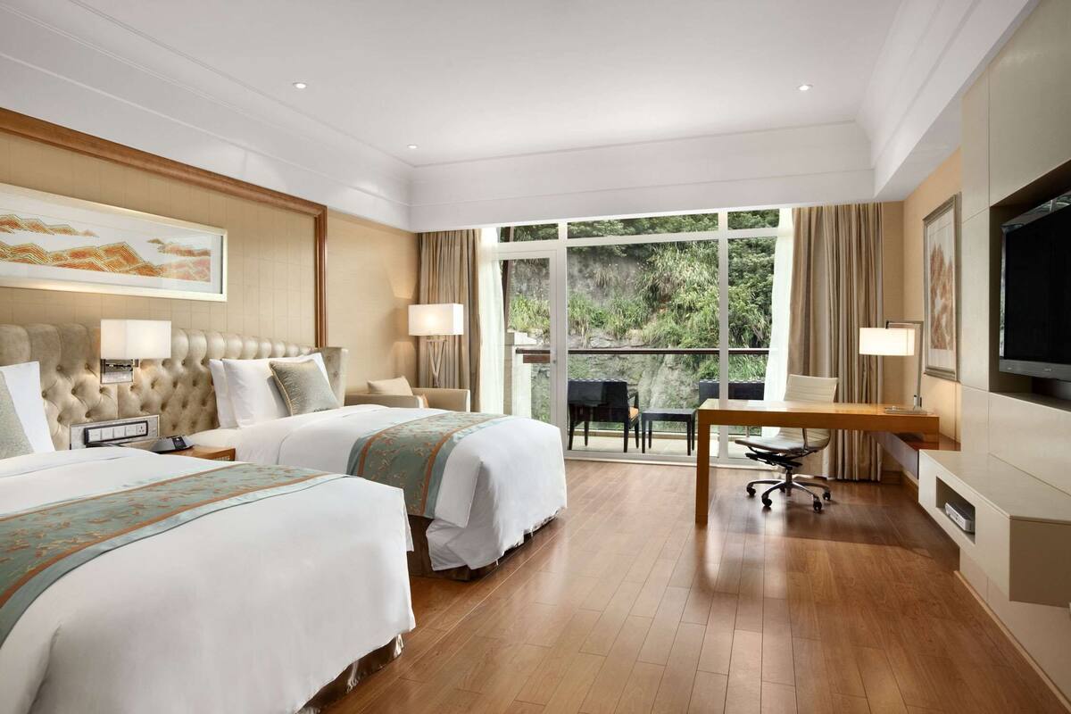 A hotel room with two beds, a desk, a TV, and a balcony with a view of greenery.