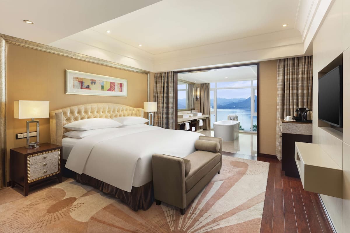 A hotel room with a large bed, a desk with a flat-screen TV, a bathroom with a view, and a balcony.