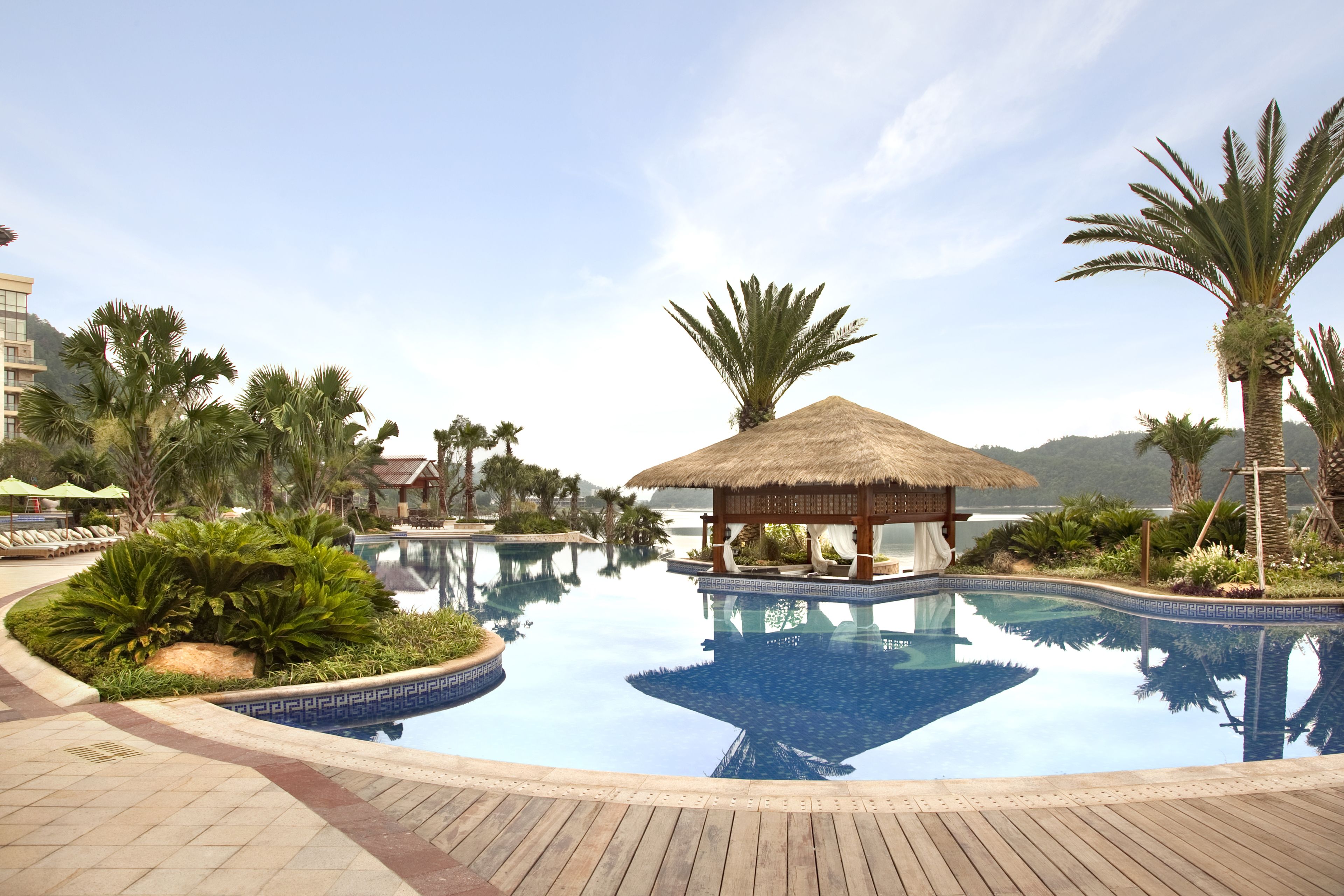 A serene hotel pool area with a thatched-roof gazebo, palm trees, and a clear blue sky.