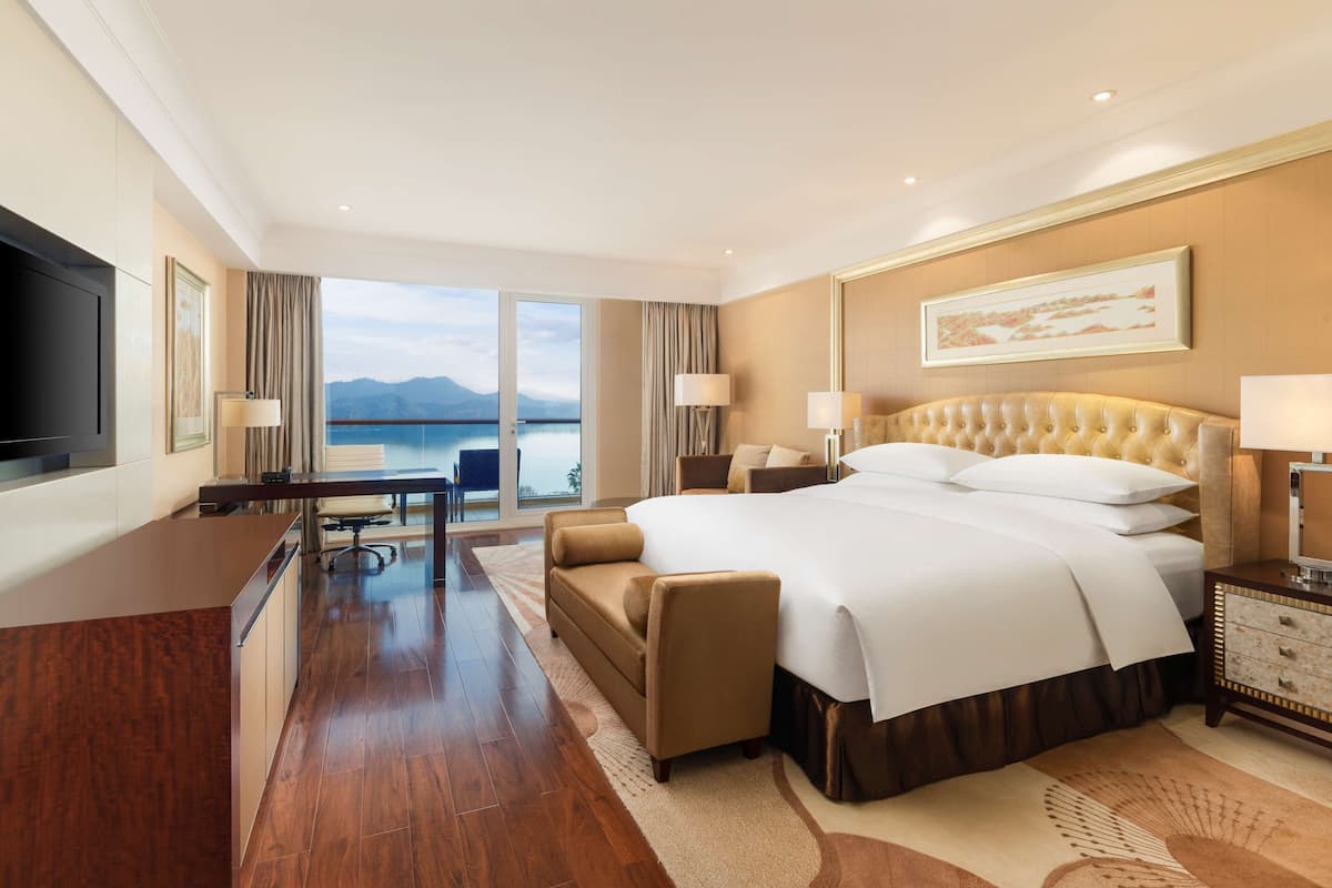 A hotel room with a large bed, a desk, a chair, a TV, and a view of the sea.