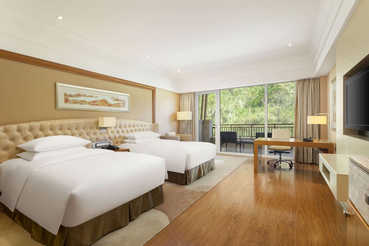 A hotel room with two beds, a desk, a TV, and a balcony with a view of greenery.