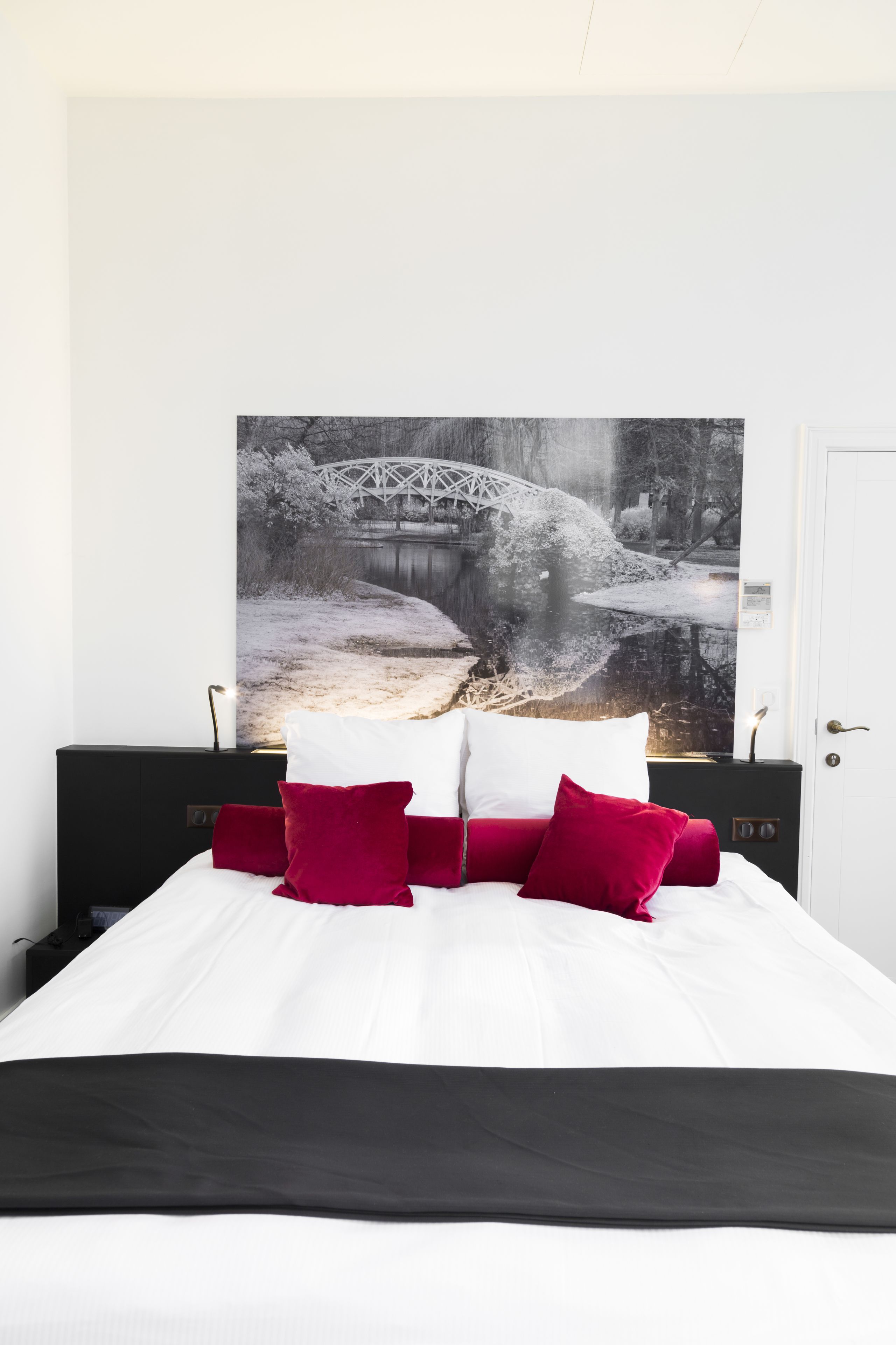 A bed with white bedding and red pillows, a black headboard, and a large black-and-white photograph of a bridge over a river on the wall behind it.