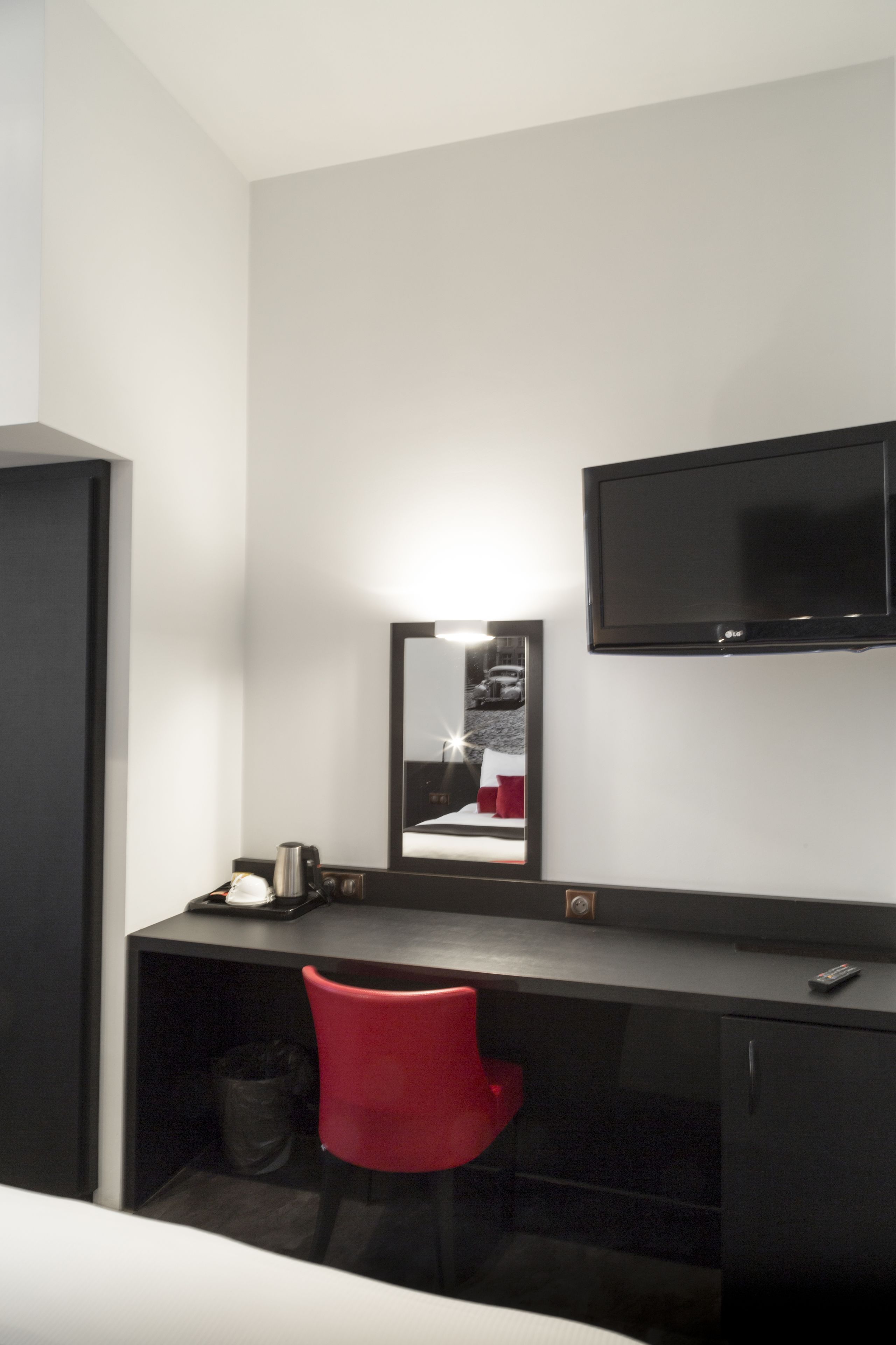Habitación de hotel con un escritorio negro, una silla roja, un televisor montado en la pared y un espejo que refleja parte de la habitación.