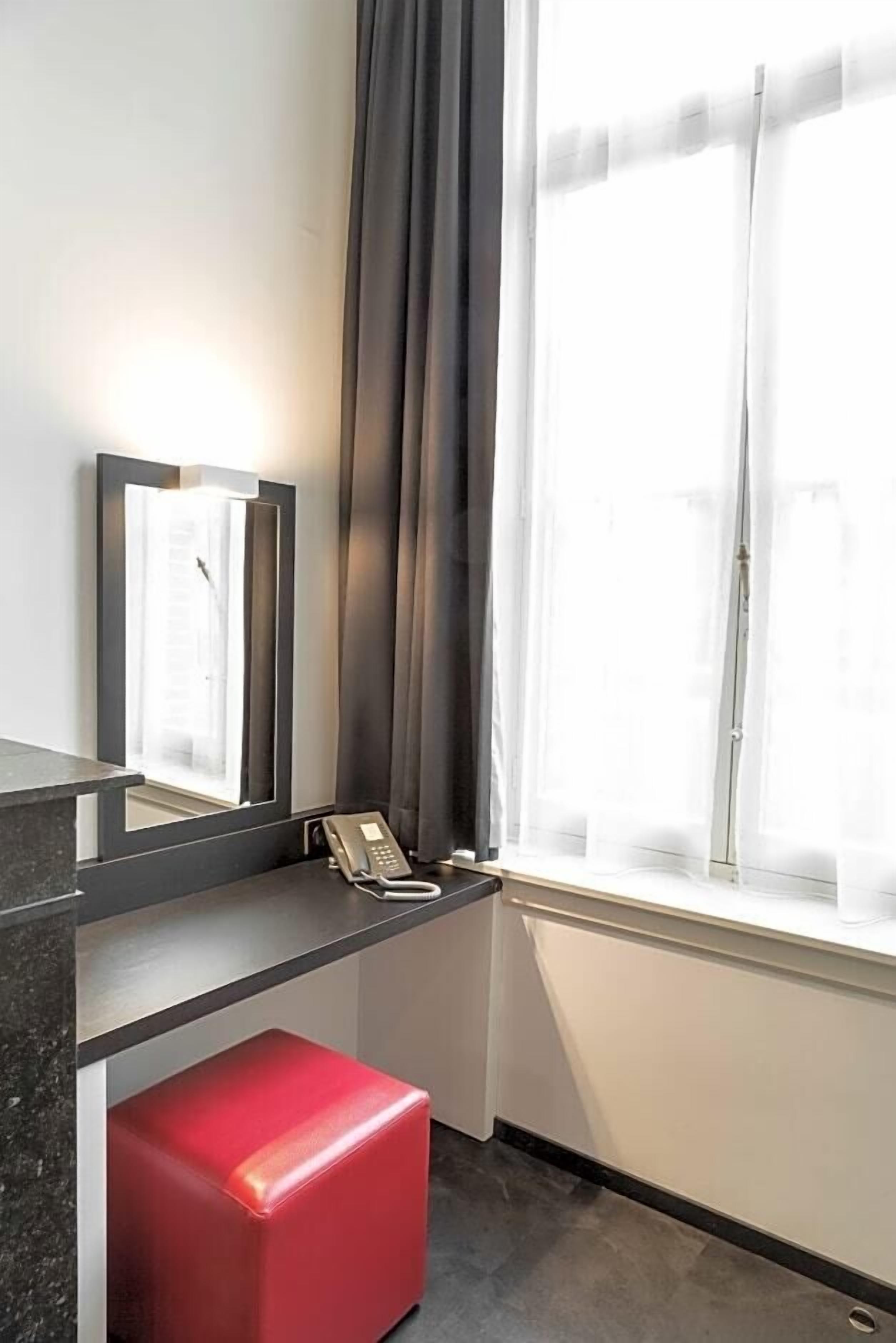 Una habitación de hotel con escritorio, teléfono, un banco rojo y una ventana con cortinas.