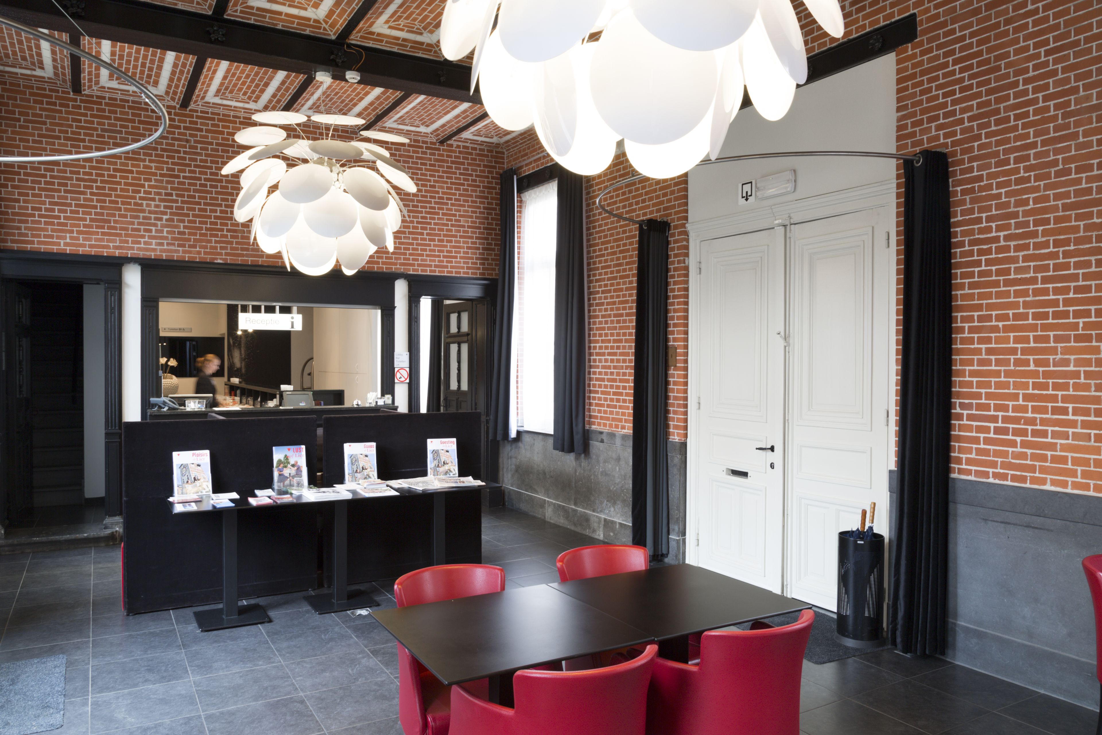 A modern interior with a brick wall, a white door, a black reception desk, and red chairs around a table.