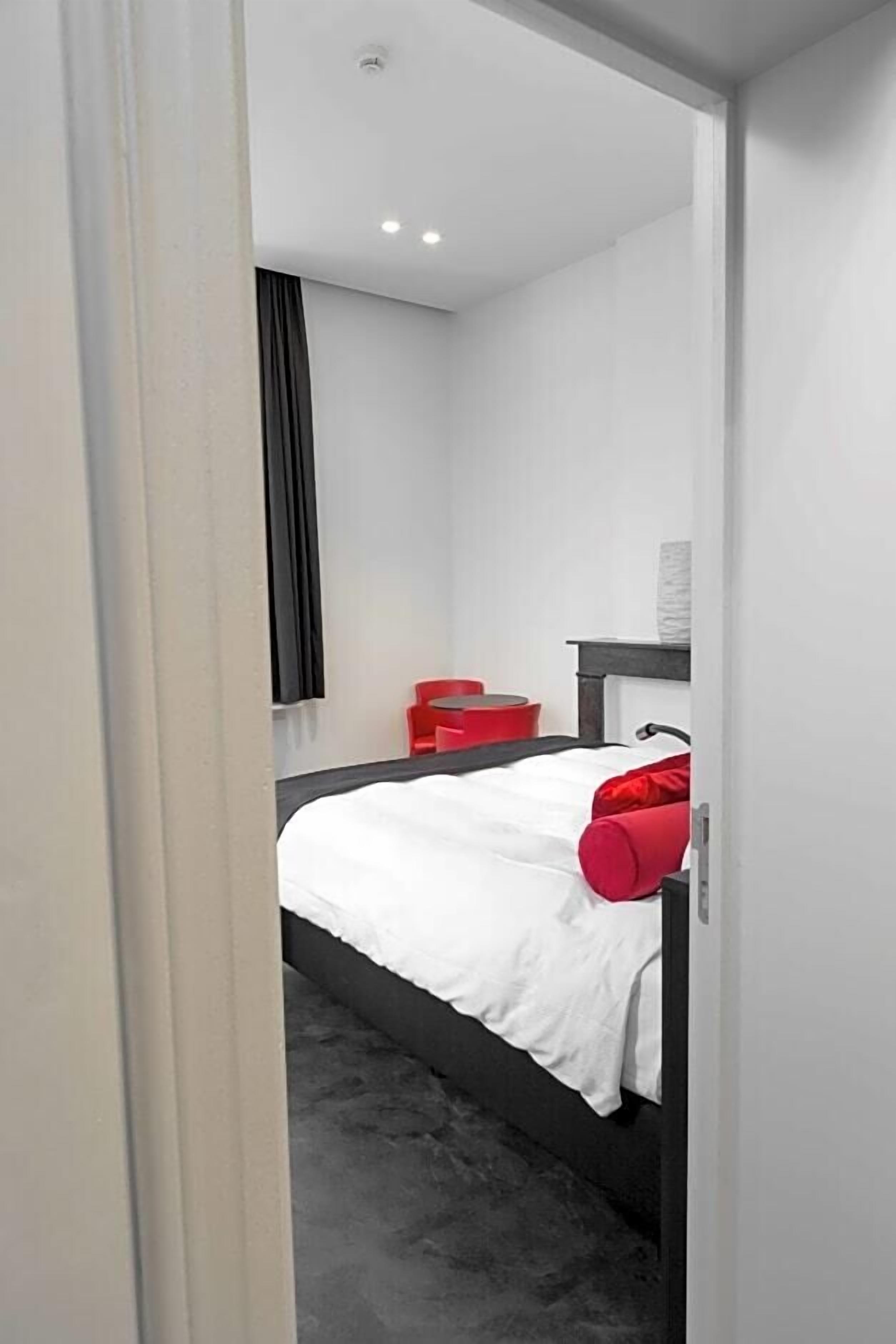 Une chambre d’hôtel moderne avec un lit, un fauteuil rouge, une petite table et une fenêtre avec des rideaux.
