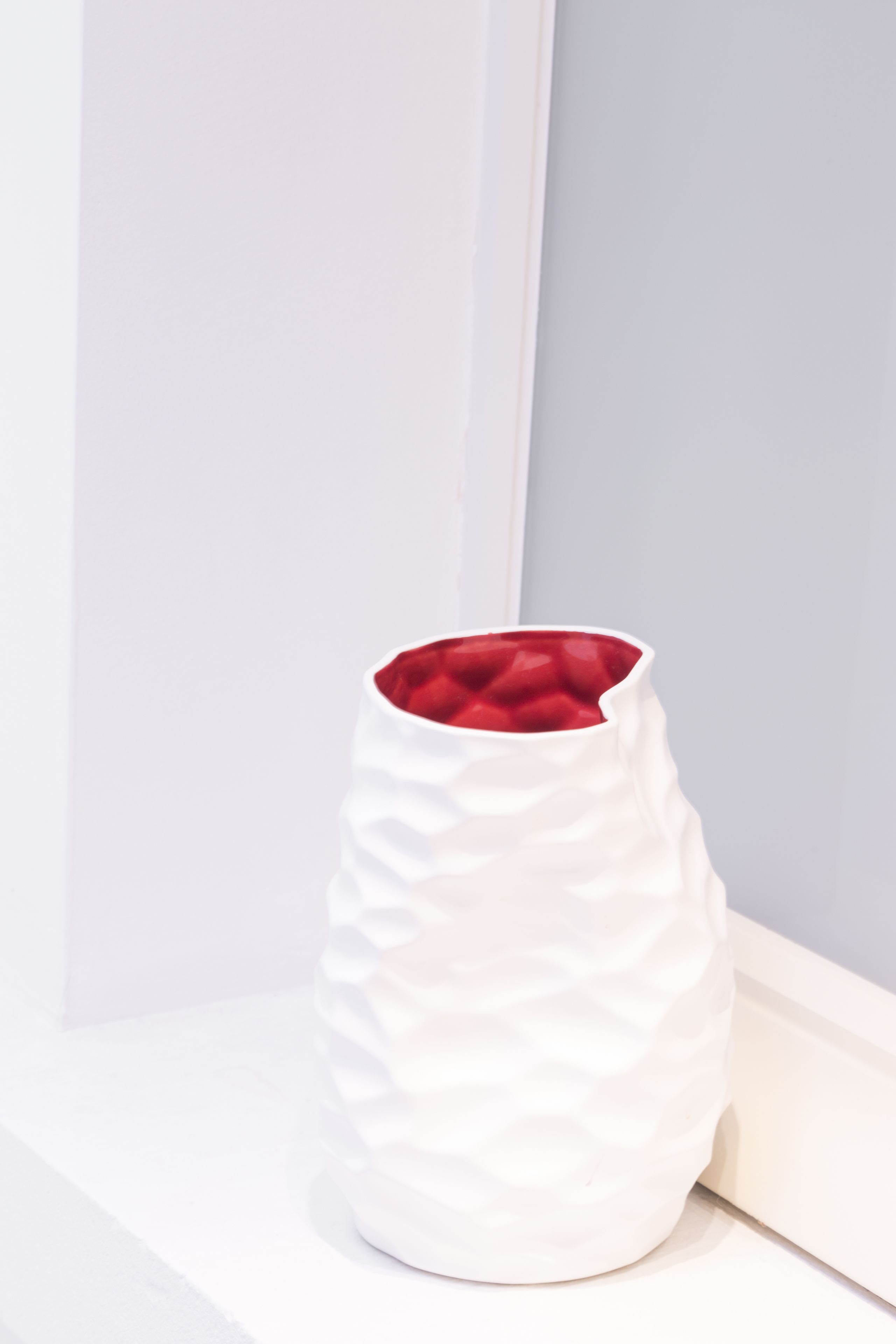 A white vase with a textured exterior and a red interior, placed on a white surface against a light-colored wall.