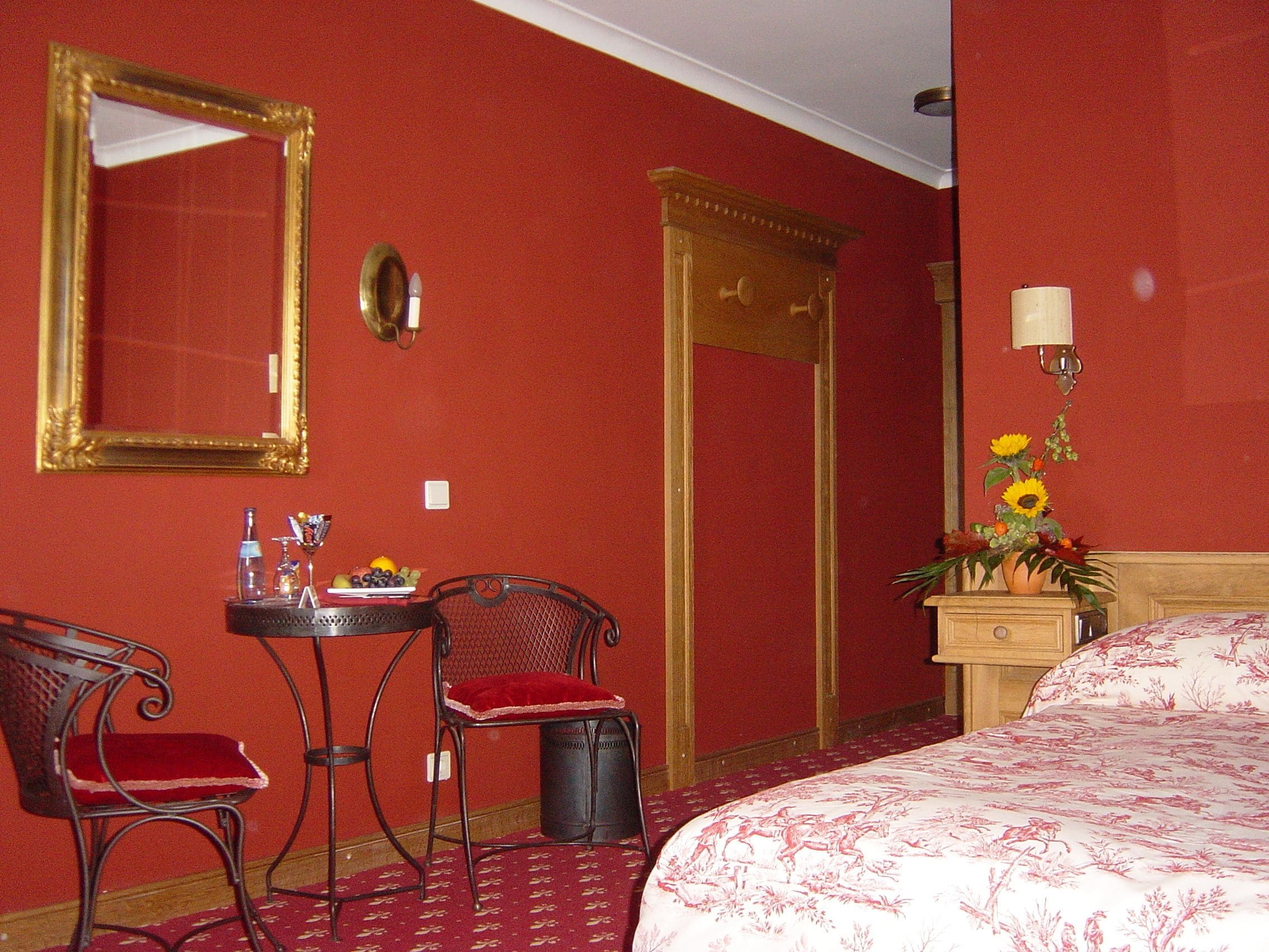 A hotel room with a bed, a desk with a chair, a mirror, a wall clock, and a nightstand with a lamp and flowers.