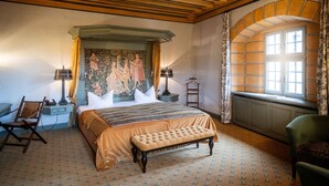 A bedroom with a large bed, a bench, a chair, a nightstand, a lamp, a tapestry, and a window with curtains. - Burg Rabenstein (Ahorntal)