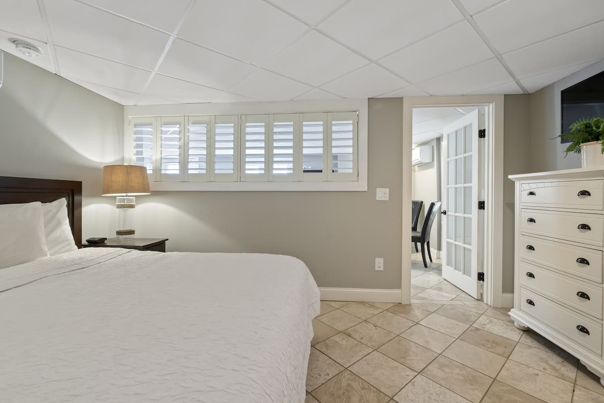 A bedroom with a bed, a nightstand, a lamp, a dresser, and a window with blinds.