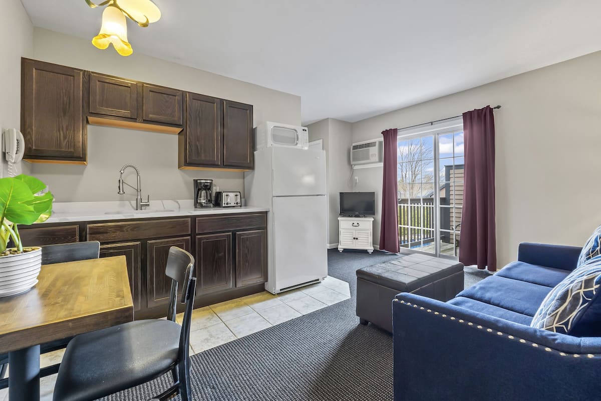 A compact living space with a kitchenette, a dining area, a sofa, and a sliding glass door to a balcony.