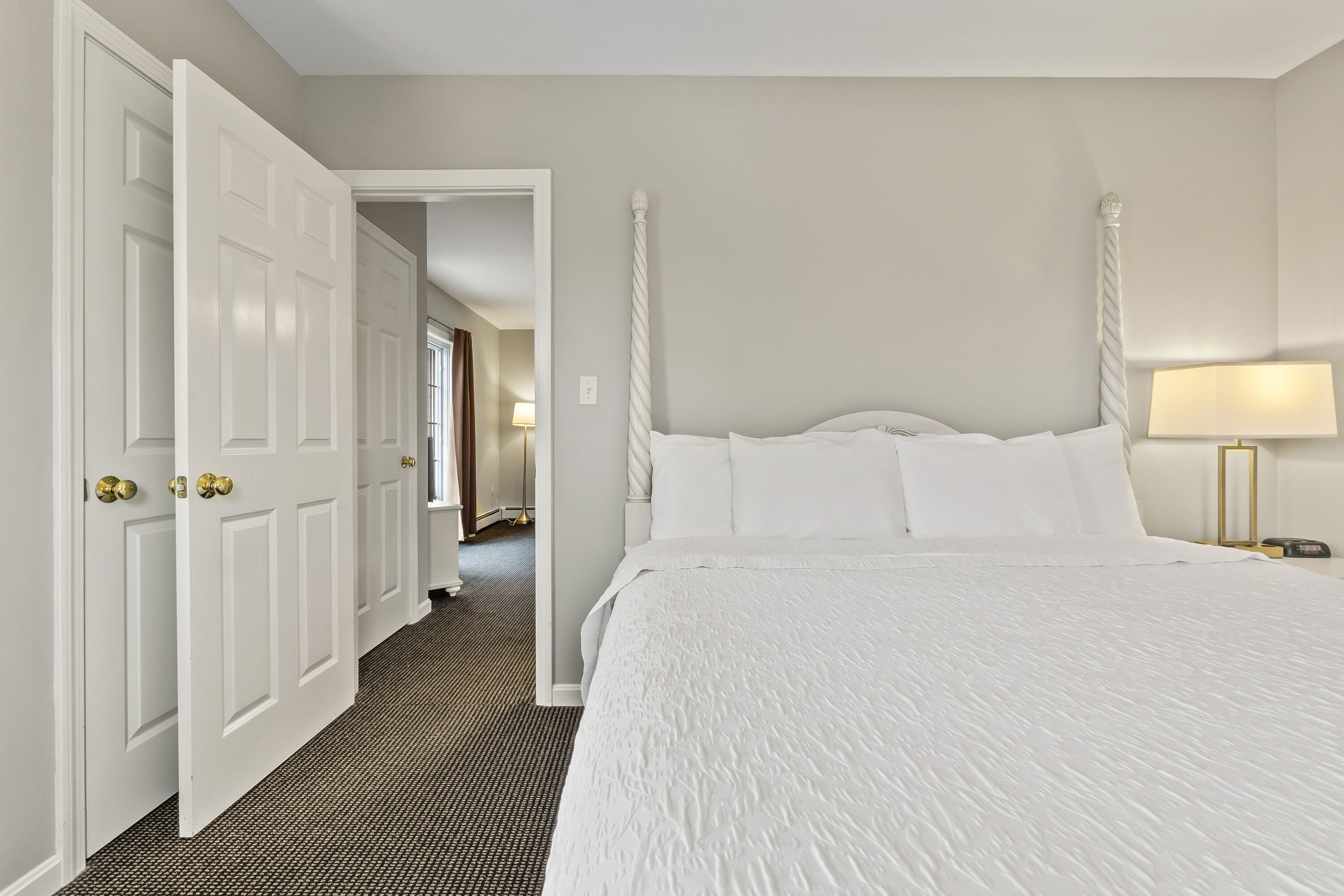 A hotel room with a large bed, a nightstand with a lamp, and a door leading to a hallway.