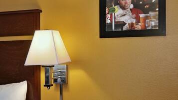 A hotel room with a bed, a nightstand with a lamp, a telephone, and a framed poster on the wall.