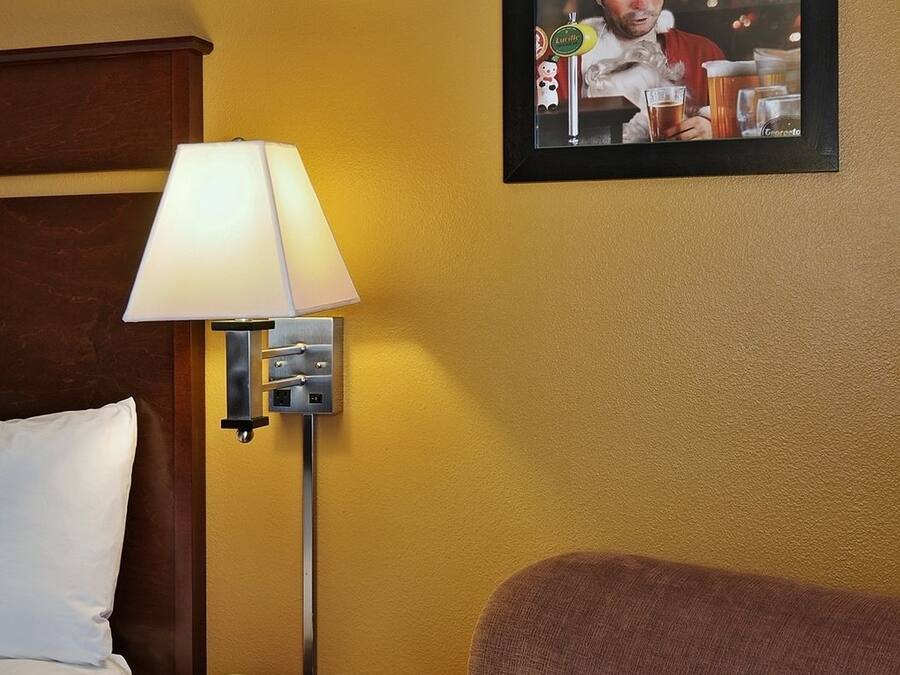 Una habitación de hotel con una cama, una mesita de noche con lámpara, un teléfono y un cartel enmarcados en la pared.