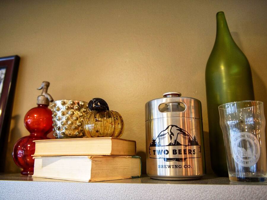 A shelf with a metal can labeled "TWO BEERS BREWING CO.", a clear glass, a green bottle, and decorative items including a red glass ornament and a wooden book.
