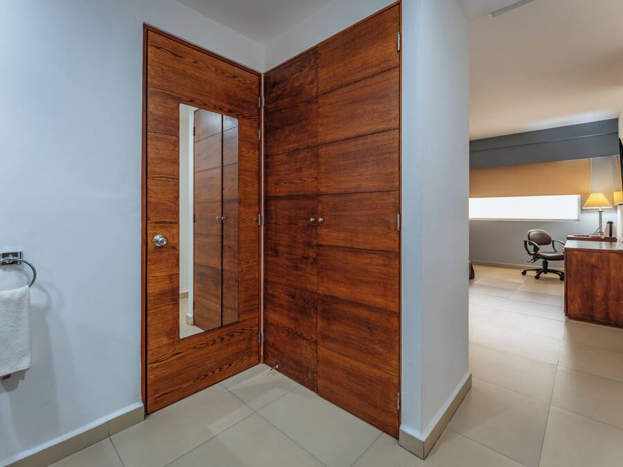 A hotel room with a wooden wardrobe, a mirror, a desk with a lamp, and a chair.