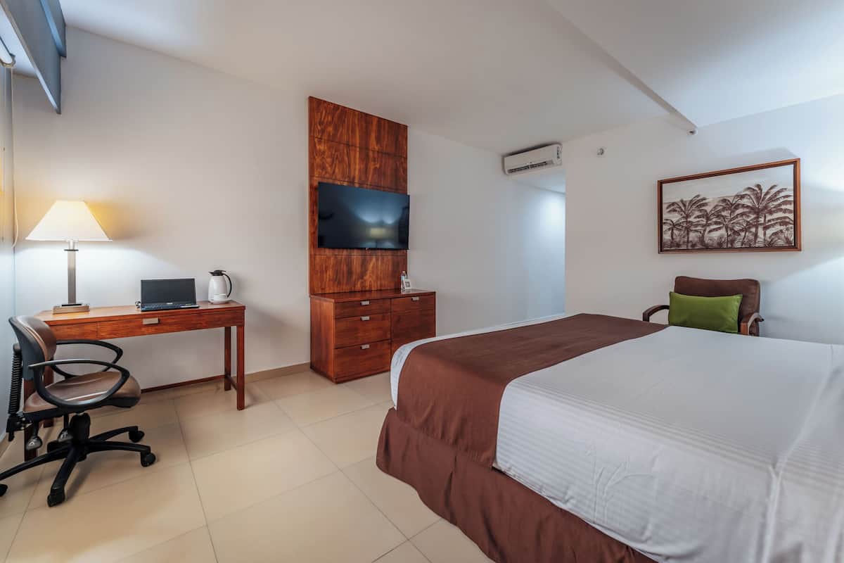 Ein Hotelzimmer mit einem Bett, einem Schreibtisch mit Computer, einem Fernseher, einem Holzschrank und einem Bild an der Wand.