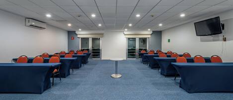 Un salón de conferencias con filas de mesas y sillas, una mesa redonda en el centro y un televisor montado en la pared.