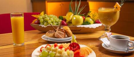 Un desayuno con una bandeja de frutas, un vaso de zumo de naranja, un plato de cruasanes y una taza de café.