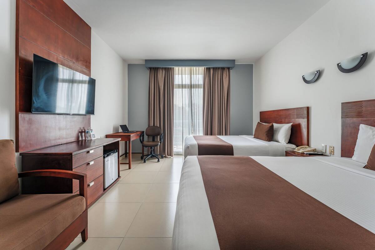Ein Hotelzimmer mit Bett, Schreibtisch, Sessel, Fernseher und einem Fenster mit Vorhängen.