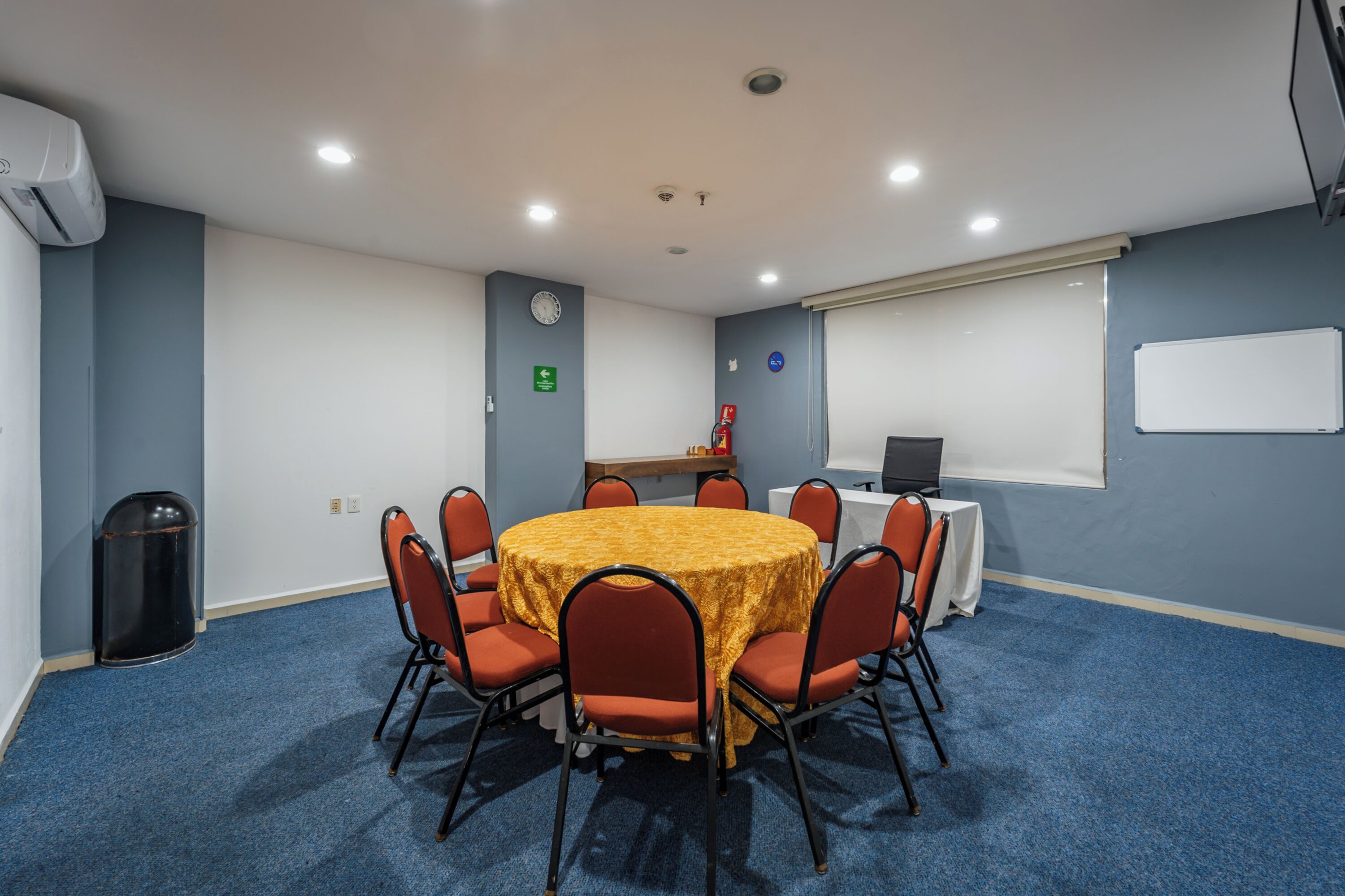 A conference room with a round table, chairs, a clock, a fire extinguisher, and a whiteboard.