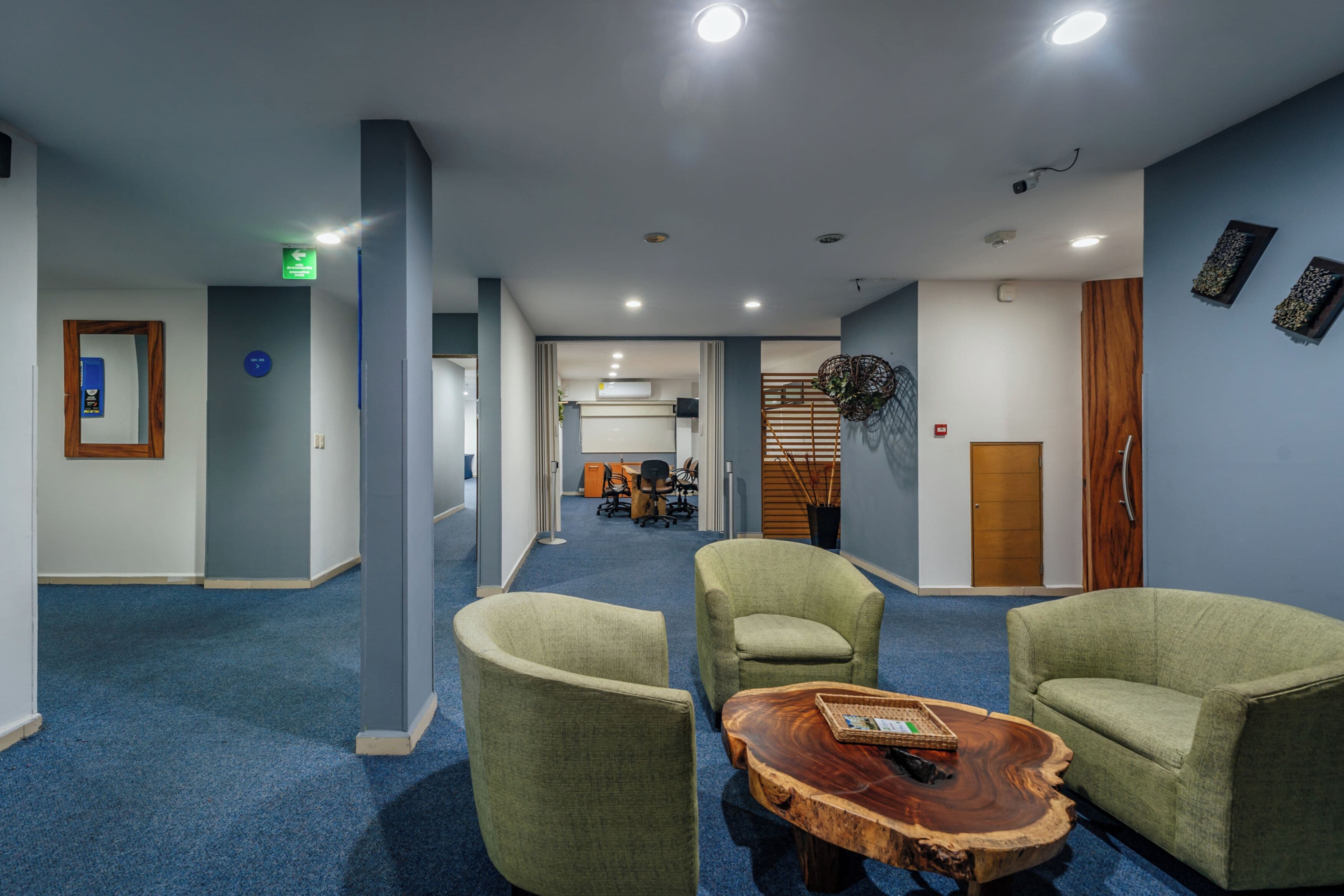 A modern hotel lobby with a blue carpet, green upholstered chairs, a wooden coffee table, and a reception area in the background.