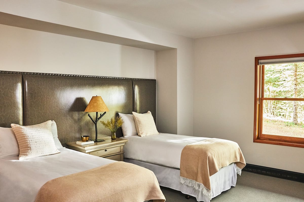 A hotel room with two beds, a nightstand with a lamp, a window with a view of greenery, and a wooden headboard.