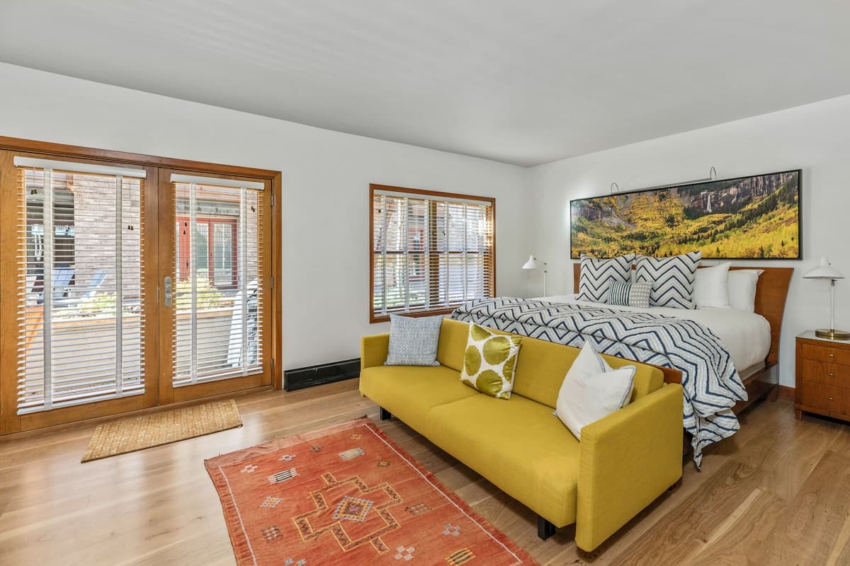 A bedroom with a bed, a yellow sofa, a red rug, and two windows with blinds.