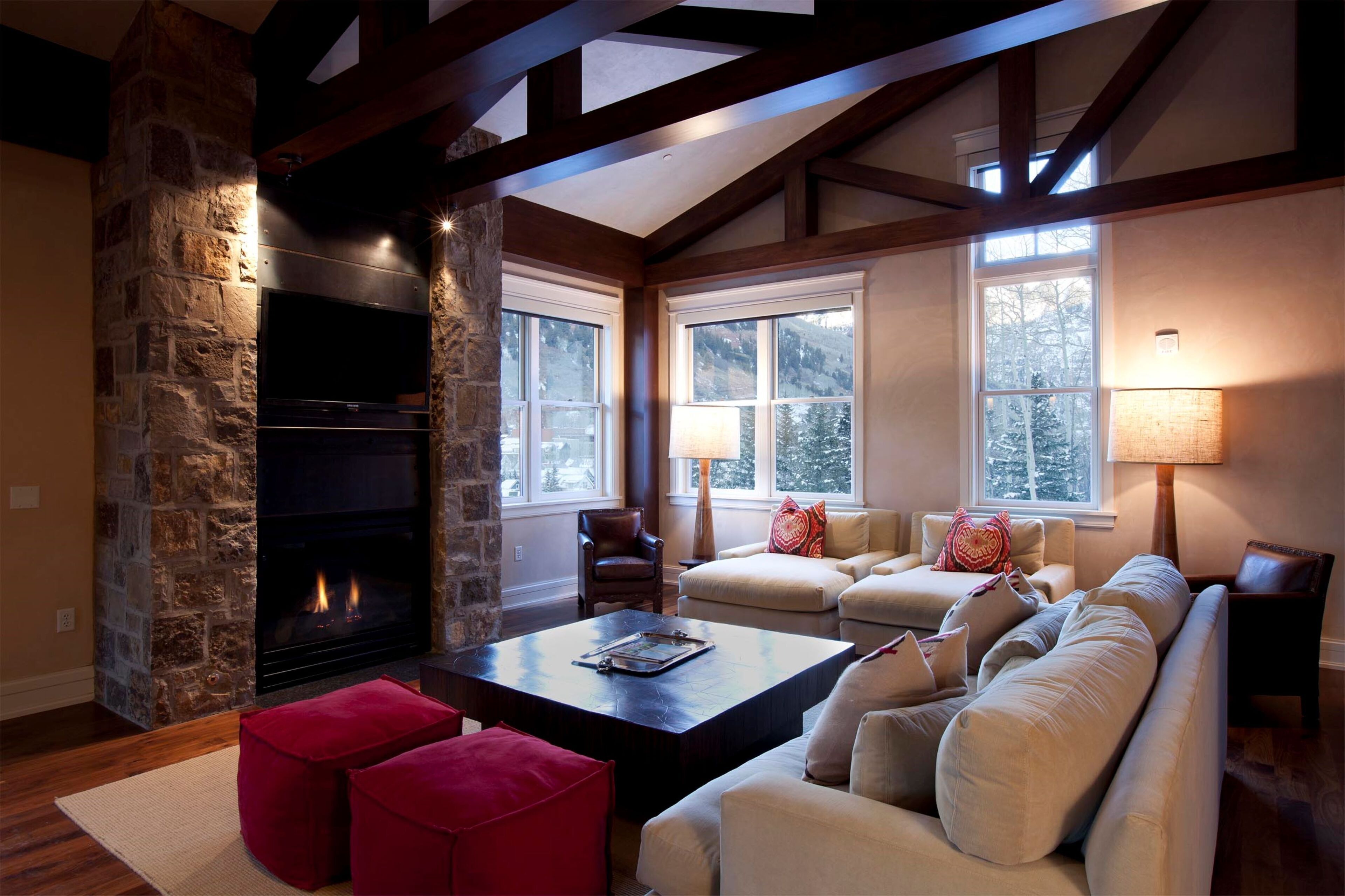 A cozy living room with a stone fireplace, wooden beams, large windows, and a comfortable seating area.