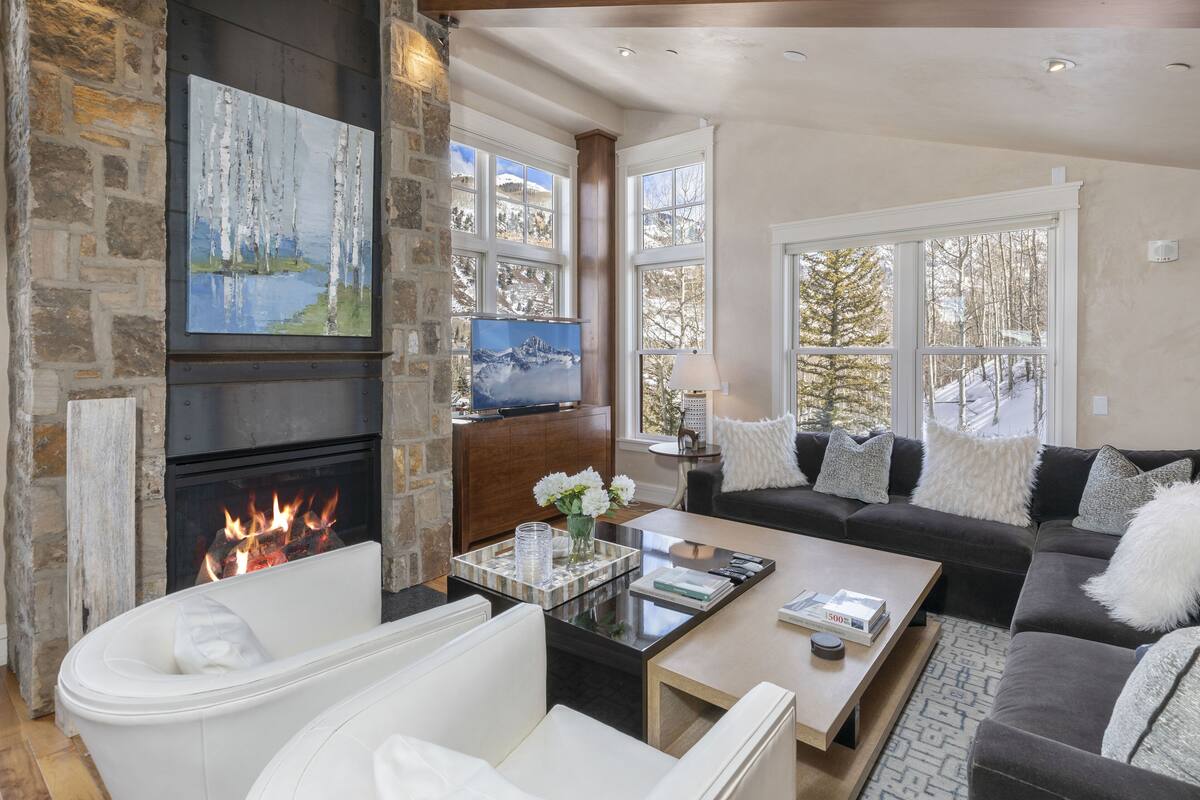 A cozy living room with a stone fireplace, a large window with a view of trees, a comfortable sofa, and a coffee table with books and a vase of flowers.