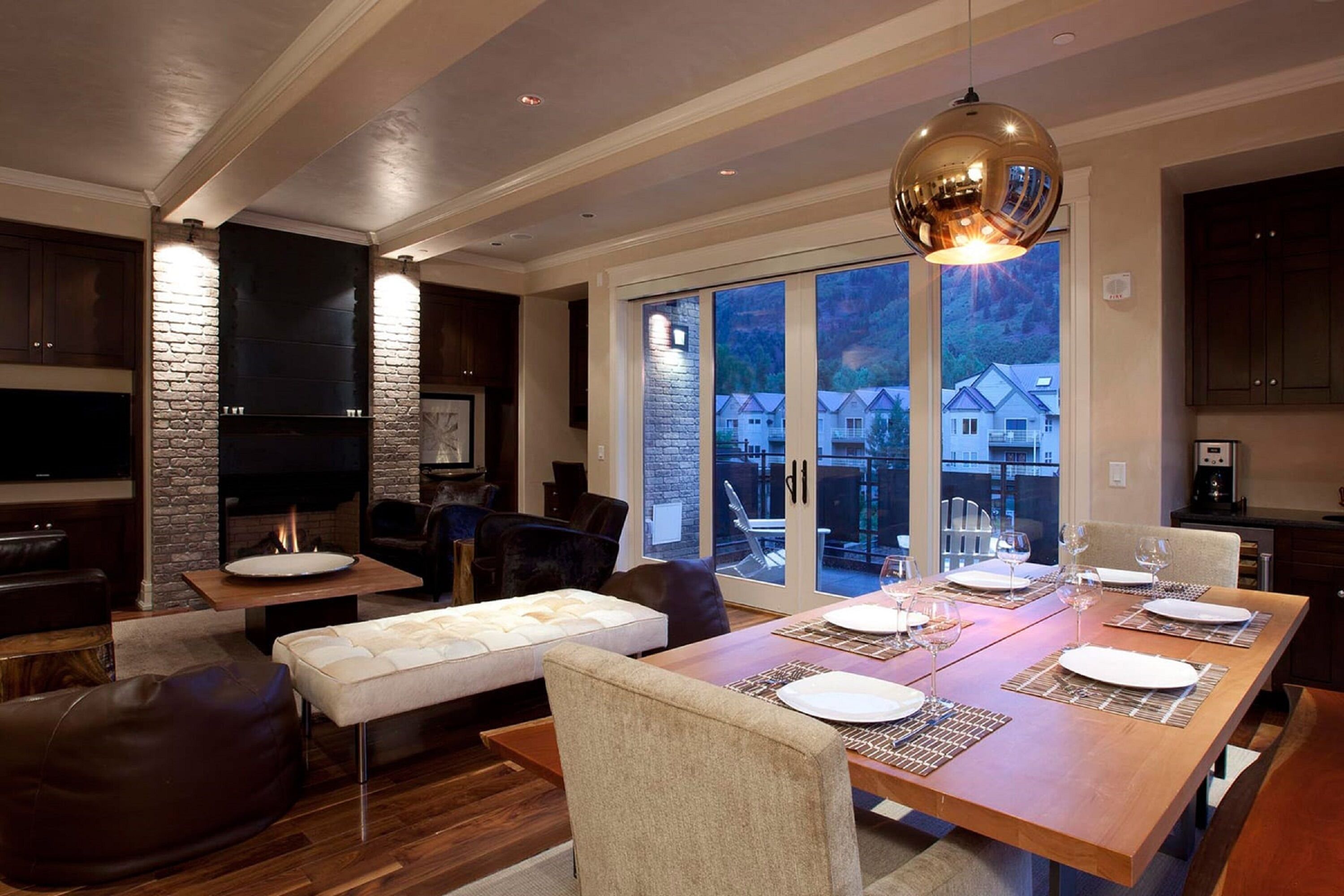 A modern dining area with a wooden table set for four, a fireplace, and a large window overlooking a residential area.