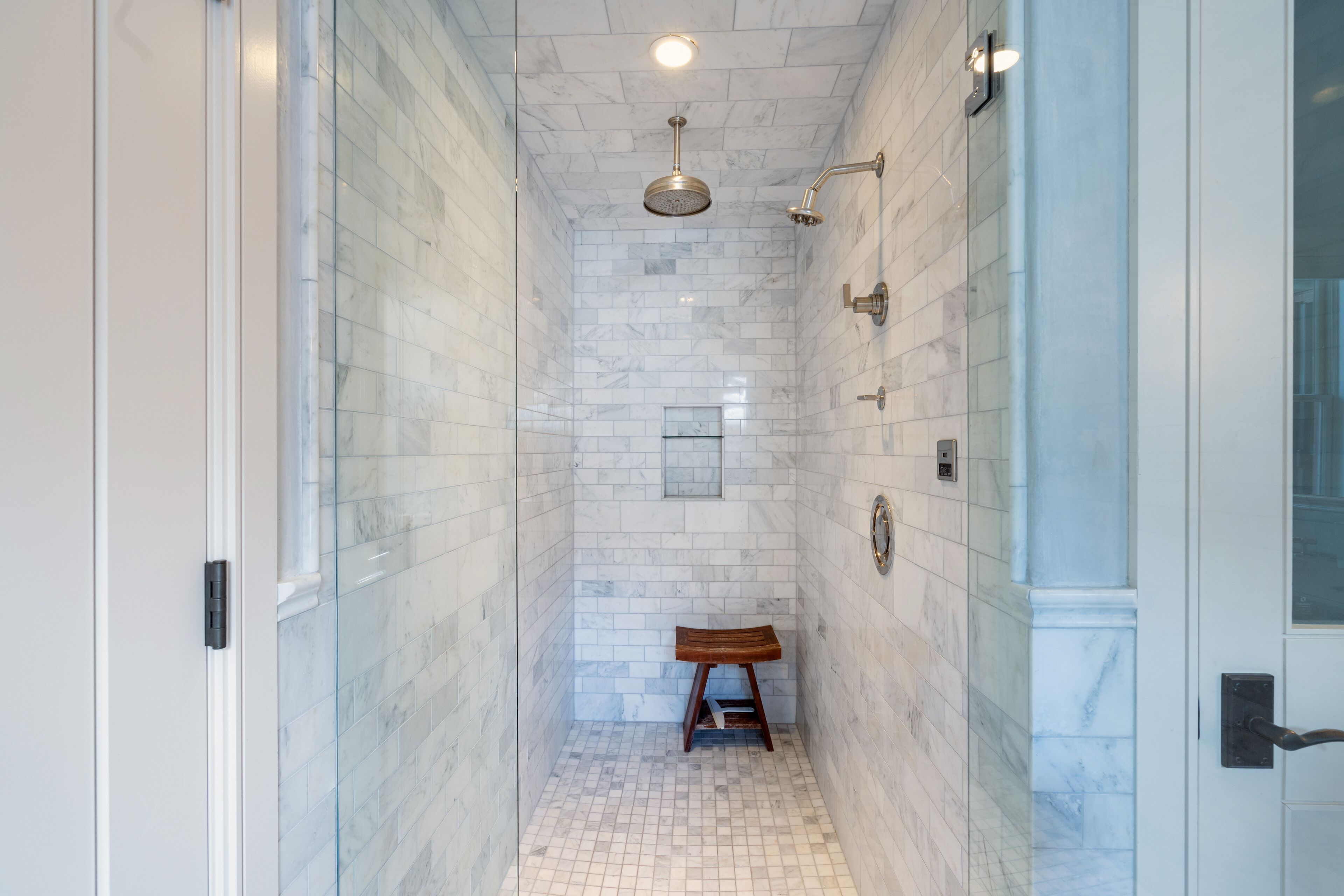 A narrow, tiled shower with a wooden stool, a handheld showerhead, and a wall-mounted control panel.