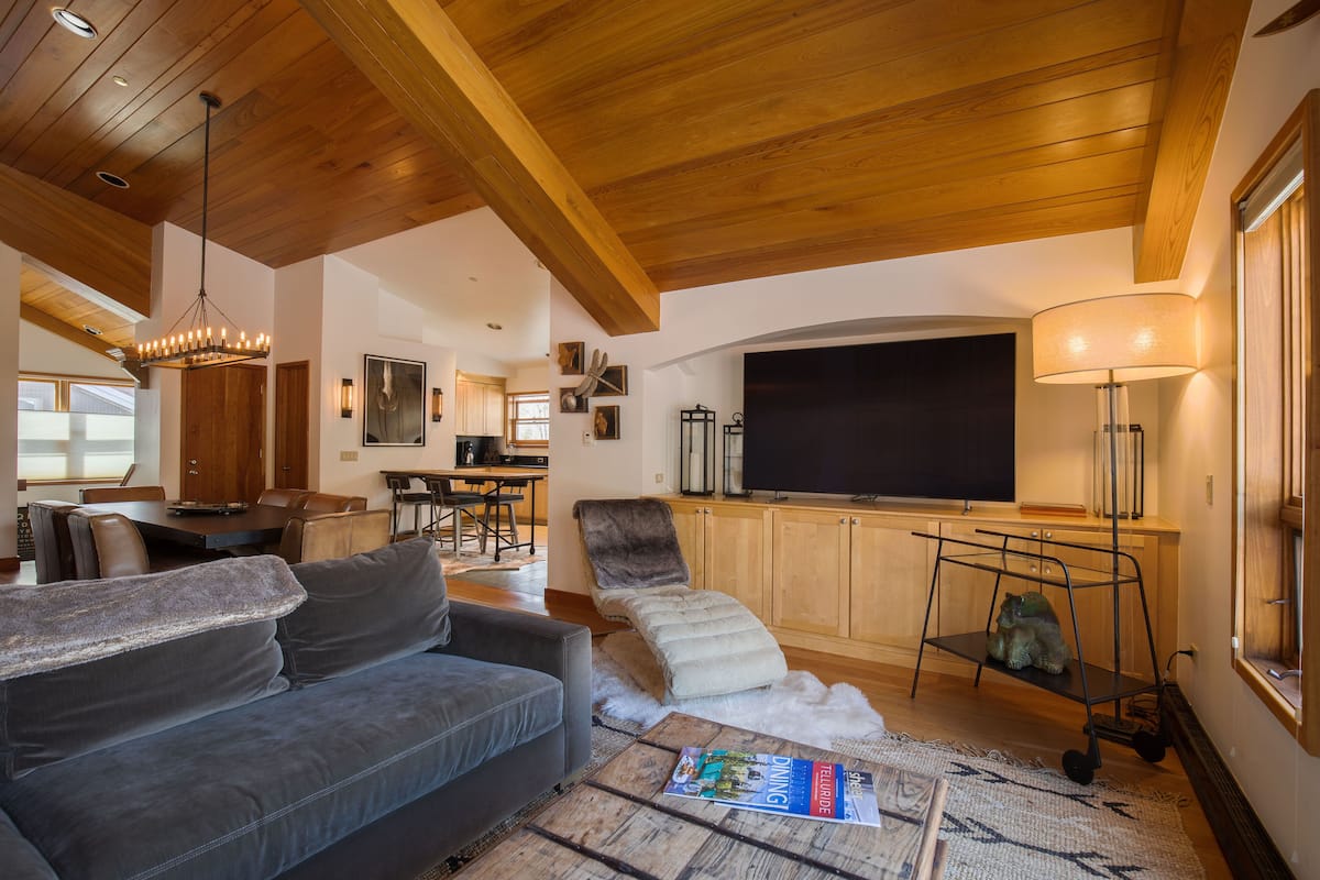 A spacious living area with a wooden ceiling, a gray sofa, a white pouf, a coffee table with a magazine, and a TV against the wall.