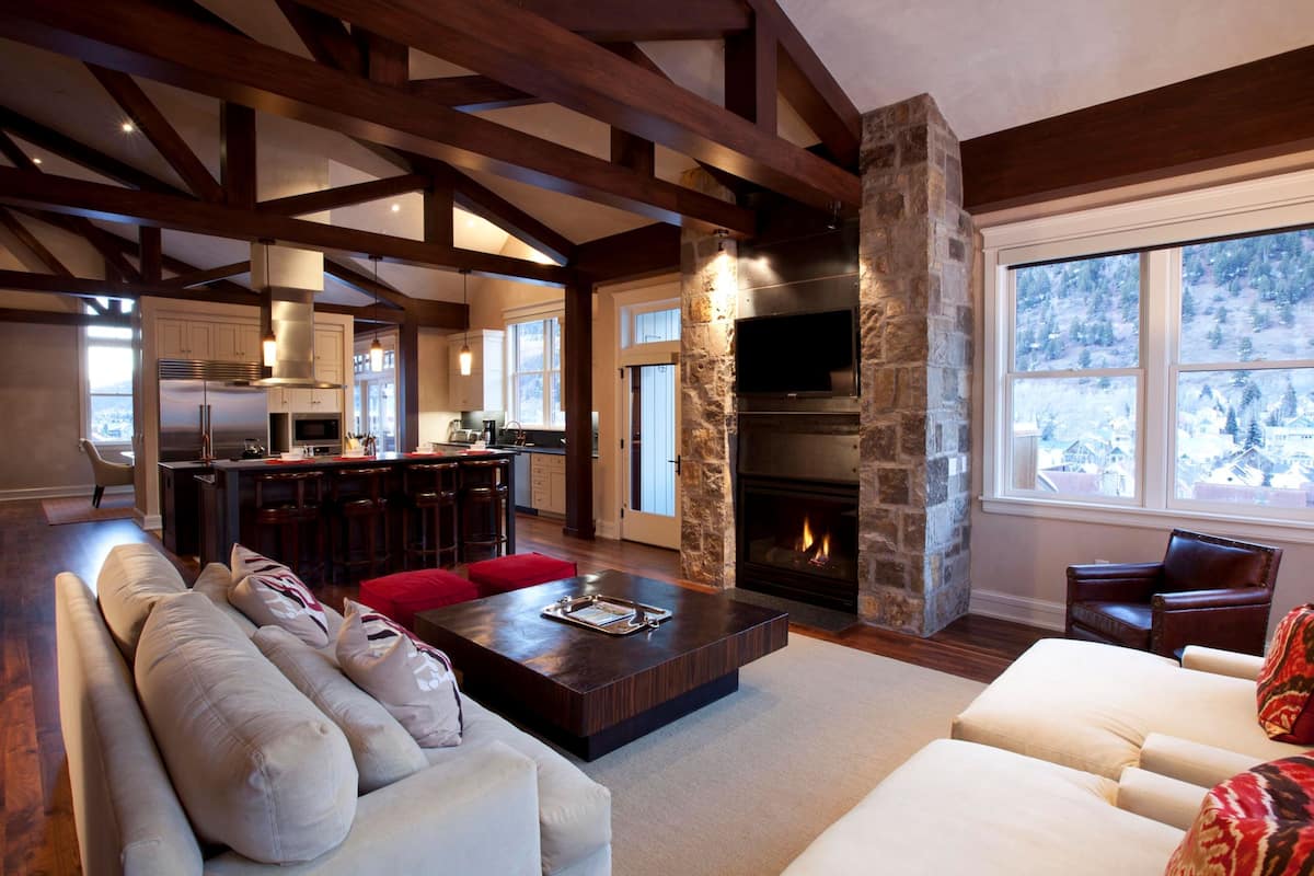 A spacious living area with a stone fireplace, wooden beams, a sofa, and a view of a snowy landscape through large windows.