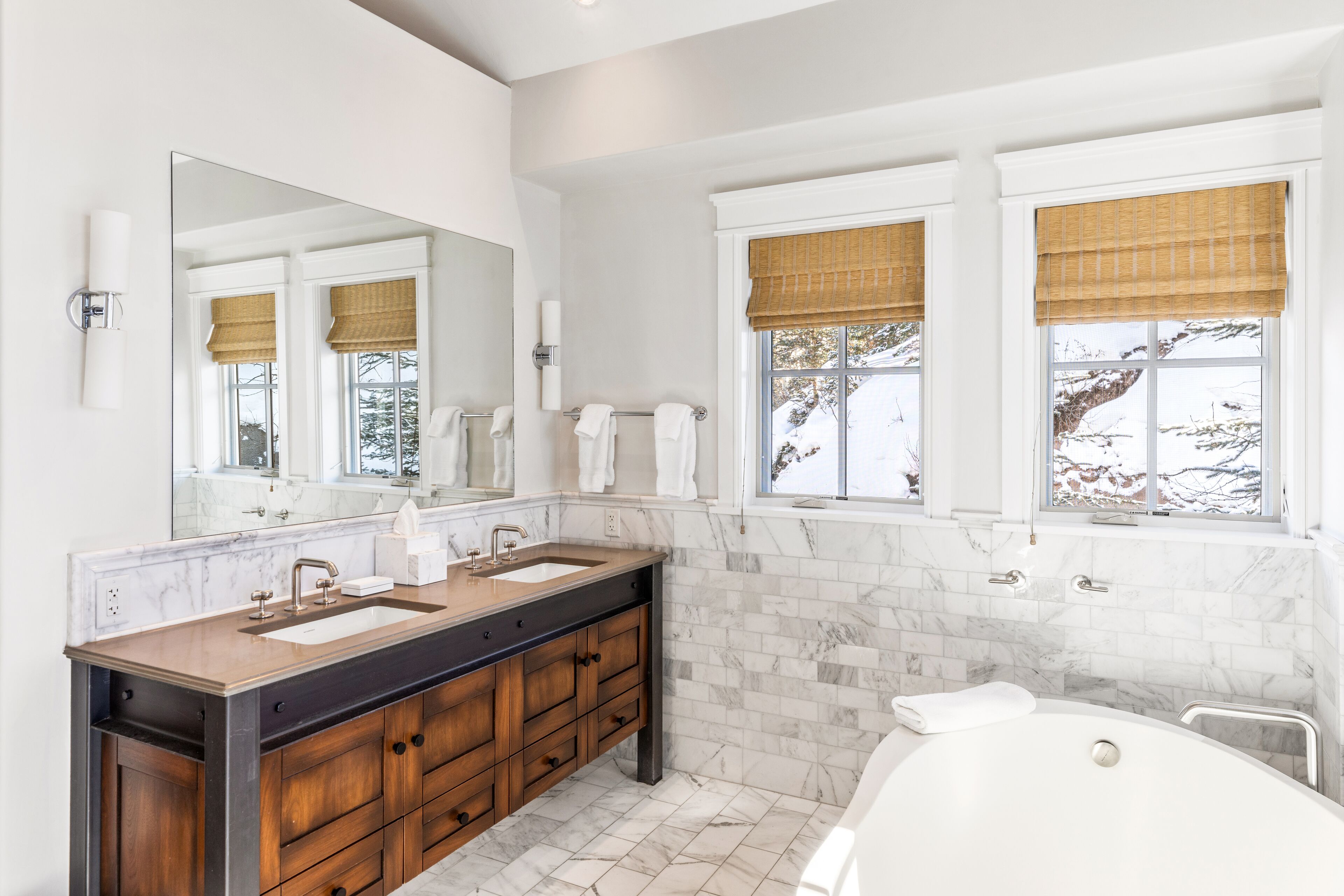 A modern bathroom with a large mirror, two sinks, a bathtub, and a window with a view of trees.